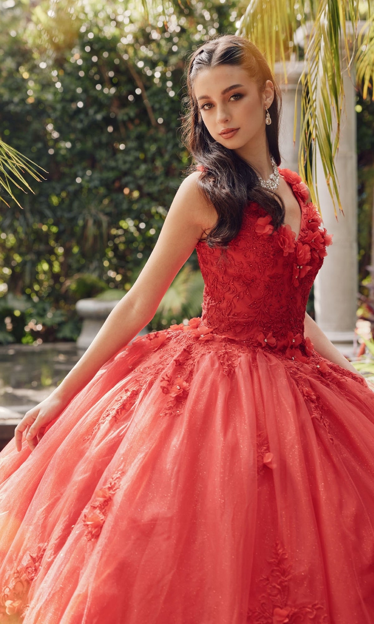 A young woman wearing the Juliet 1437R Red Ball Gown Quinceañera Dress with beaded embroidery poses outdoors among lush greenery, gazing at the camera.